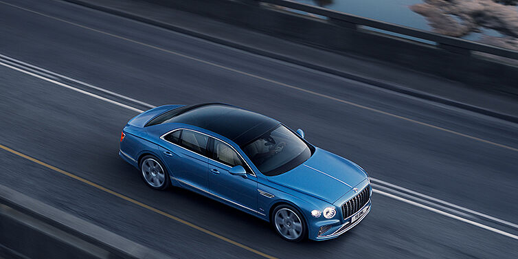 Bentley Knokke New Bentley Flying Spur Azure sedan in Light windsor blue paint by Mulliner, with 22 inch Azure wheels seen from overhead driving dynamically on a country road